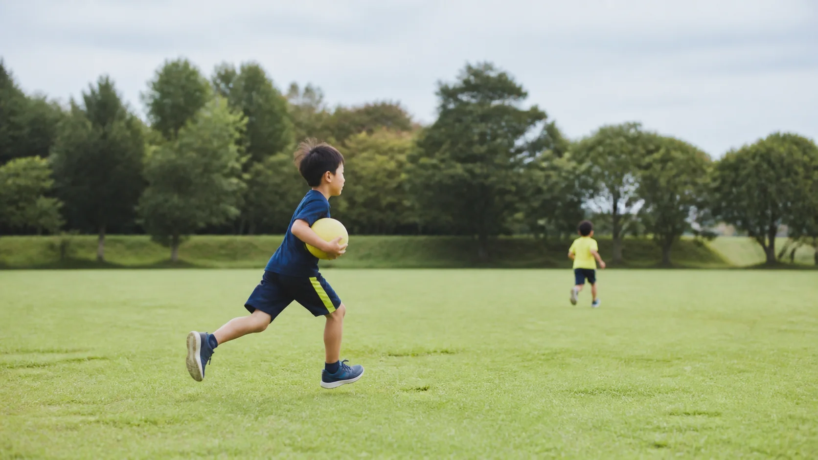 芝生の上をボールを持って走る日本人の子ども